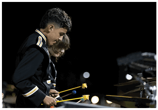 Two people in dark uniforms are playing percussion instruments with yellow mallets in a dimly lit outdoor setting. Description generated by AI