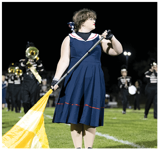 A person wearing a navy blue dress with a white collar and holding a yellow flag stands on a grassy field with a marching band playing instruments in the background at night. Description generated by AI
