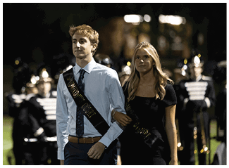Two adults dressed formally with sashes walk arm in arm on a field at night with a marching band in uniform behind them. Description generated by AI