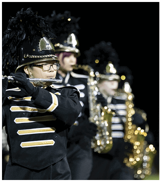 People in marching band uniforms are playing wind instruments in a line outdoors at night. Description generated by AI