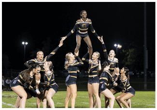 A group of people in matching black and gold cheerleading uniforms perform a stunt on a grassy field at night. Description generated by AI
