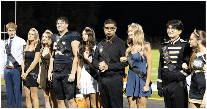 Five people wearing formal and semi-formal attire with sashes and tiaras stand together in an outdoor nighttime setting. Description generated by AI