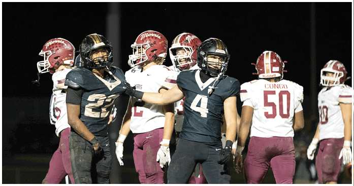 Two people in black football uniforms are interacting on the field while others in white and maroon uniforms stand in the background. Description generated by AI