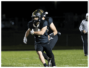 Two people in football uniforms and helmets are running on a field at night, with one person holding a football securely. Description generated by AI