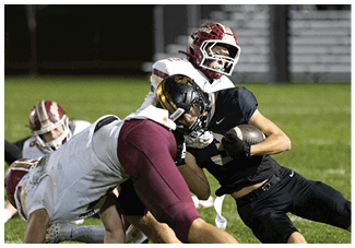 Two people wearing football helmets and uniforms are engaged in a tackle during a game on a grassy field with bleachers in the background. Description generated by AI