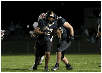 A person in a black football uniform is running with the ball while being pursued by another player in a white and red uniform during a nighttime game. Description generated by AI
