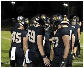 Two people in black football uniforms and helmets with gold logos stand closely facing each other on a field at night. Description generated by AI