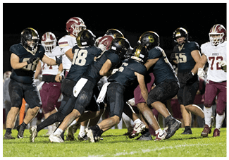 A group of people wearing dark and maroon football uniforms are engaged in a tackle on a grassy field during a nighttime game. Description generated by AI