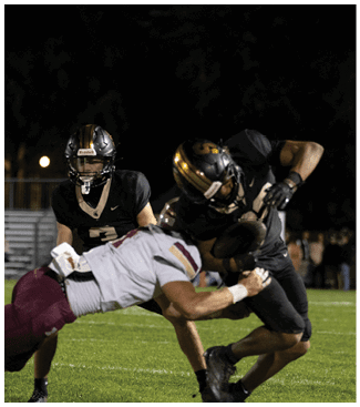 Two people in football uniforms are engaged in a tackle on a grassy field during a nighttime game. Description generated by AI
