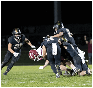 Four people in football uniforms are engaged in a tackle on a grassy field under stadium lights at night. Description generated by AI