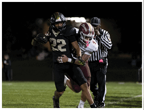 A person wearing a black football uniform with the number 22 is running with the ball while another person in a white and maroon football uniform attempts to tackle them on a grassy field at night. Description generated by AI