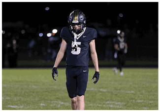 A person wearing a black football uniform with the number five stands on a field at night with another player blurred in the background. Description generated by AI