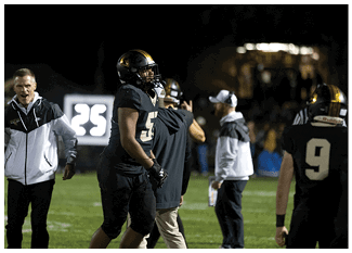 A person wearing a black football uniform and helmet stands on the sidelines of a nighttime game with two other people in the background. Description generated by AI