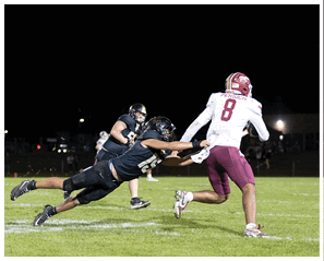 A person in a black uniform is diving and grabbing the jersey of another person in a white and maroon football uniform running with the ball on a grassy field at night. Description generated by AI