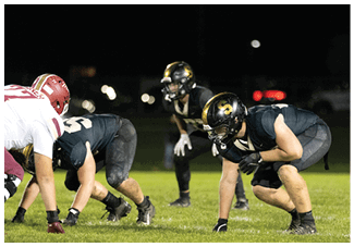 Three people in football uniforms and helmets are crouched on a grassy field at night, preparing for a play. Description generated by AI