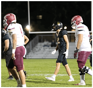 People wearing football uniforms are standing on a grassy field with bleachers and goalposts in the background during a nighttime game. Description generated by AI