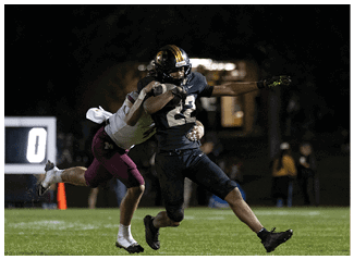 Two people in football uniforms are engaged in a tackle on a grassy field during a nighttime game. Description generated by AI