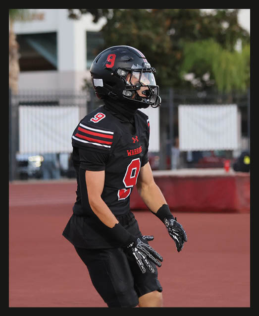 A person with curly hair wearing a black football uniform with “VISTA" on the jersey is sitting on a grassy field with one leg bent and the other extended. Description generated by AI