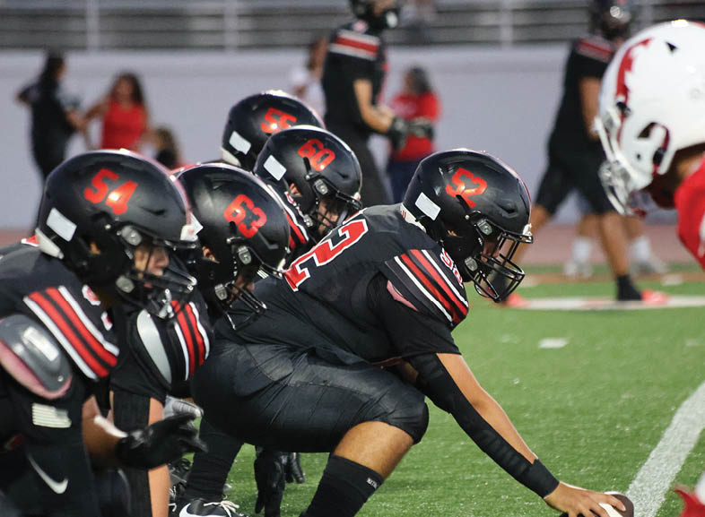 Four people wearing black football uniforms with “VISTA" on the front are standing together in a stadium with a crowd in the background, two of them holding helmets raised above their heads. Description generated by AI