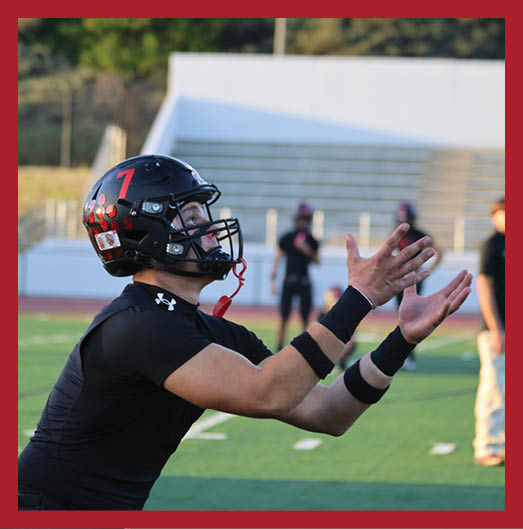A person wearing a black football uniform with “VISTA" on the jersey is sitting on a grassy field with one leg bent and the other extended, next to a black football helmet. Description generated by AI