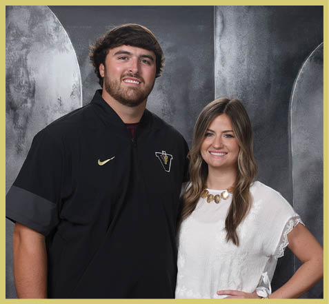 Two adults stand close together smiling in front of a gray textured backdrop, with one wearing a black sports jacket and the other wearing a white blouse with a statement necklace. Description generated by AI