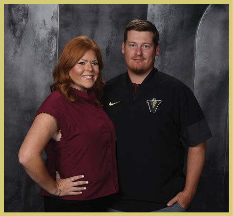 Two adults stand closely together smiling against a dark textured backdrop, with one wearing a maroon sleeveless blouse and the other wearing a black Nike jacket with a logo. Description generated by AI