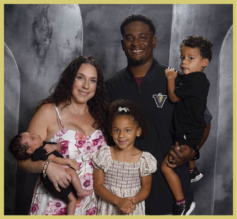 A smiling family of five poses together in front of a gray backdrop with one adult holding a newborn, another adult holding a child, and a young child standing in the center wearing a dress. Description generated by AI