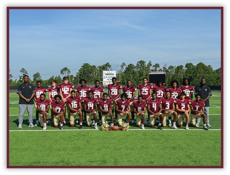 A group of people wearing maroon and gold football uniforms pose for a team photo on a green football field with two coaches standing on either side. Description generated by AI