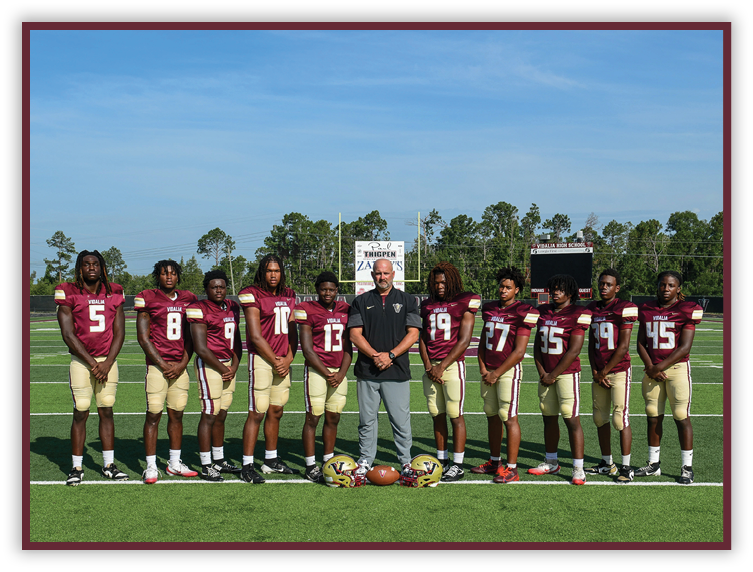 A group of people wearing maroon and gold football uniforms stand on a football field with a coach in the center holding a football. Description generated by AI
