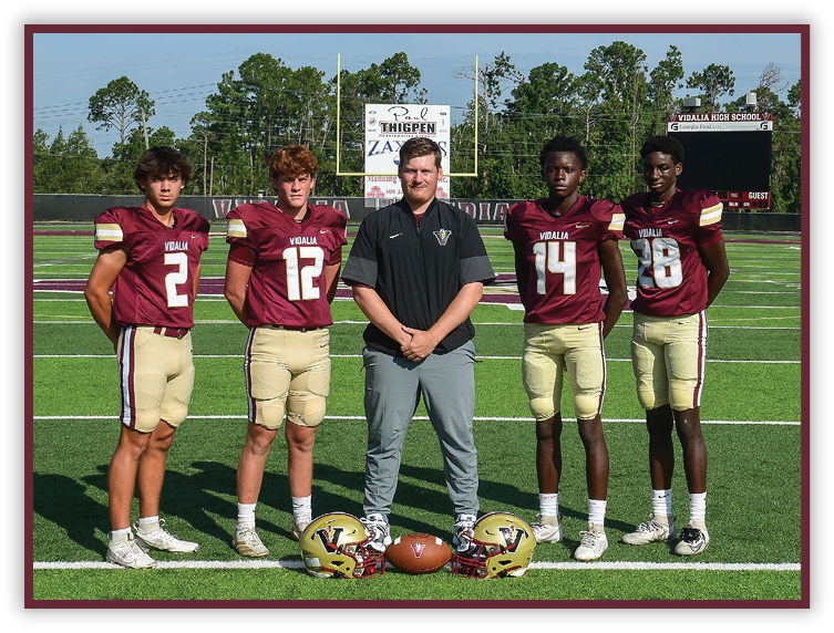 Five people wearing football uniforms and one person in casual sportswear stand on a football field with helmets and a football placed on the ground in front of them. Description generated by AI