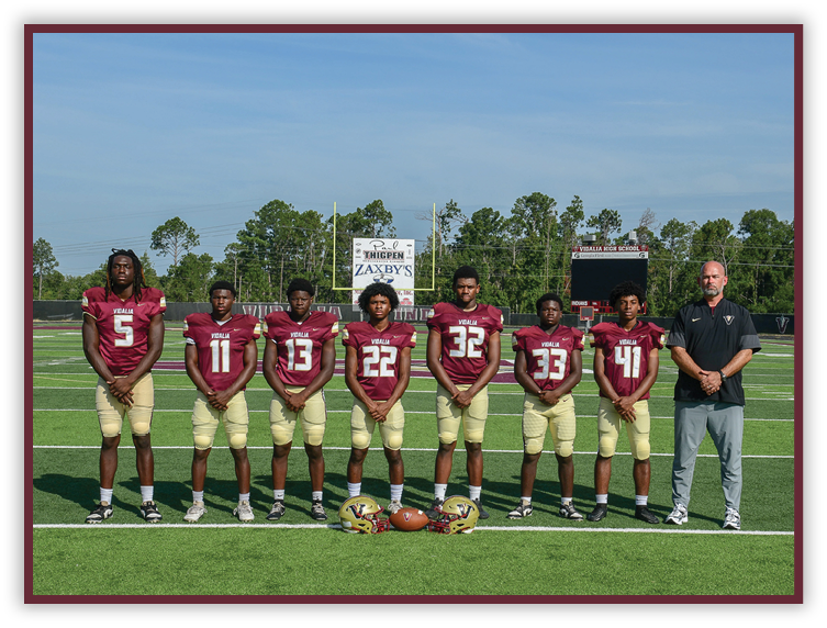 Seven people wearing matching football uniforms stand in a line on a football field with helmets and a football placed on the ground in front of them and a scoreboard visible in the background. Description generated by AI