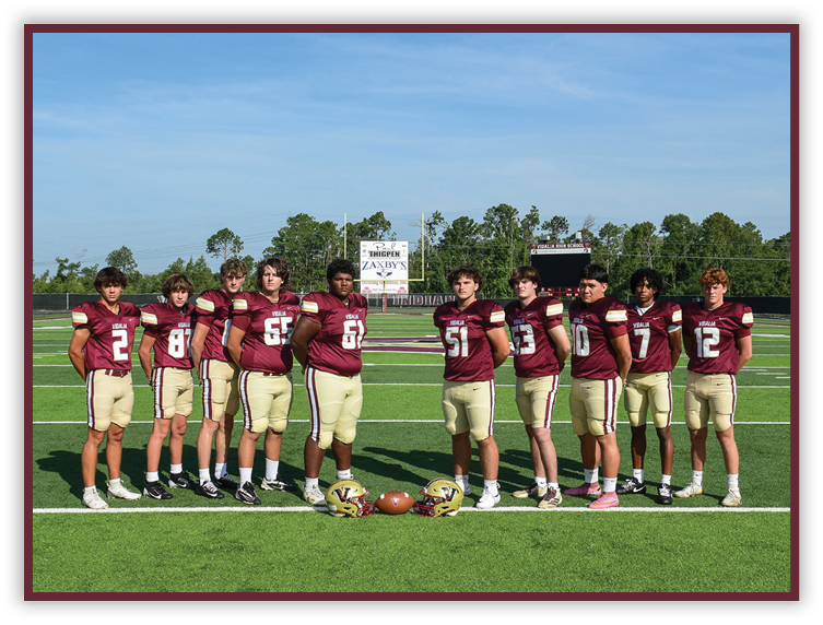 Ten people wearing maroon and gold football uniforms stand in a line on a football field with helmets and a football placed on the ground in front of them. Description generated by AI