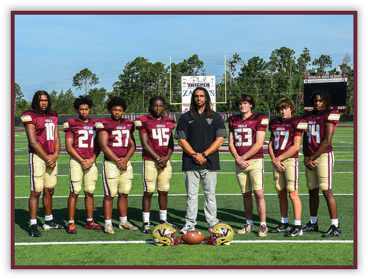 Seven people wearing matching football uniforms stand on a football field with one person in casual clothing standing in the center, and two football helmets and a football placed on the ground in front of them. Description generated by AI