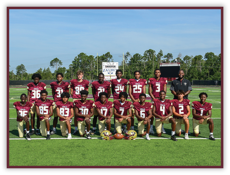 A group of people wearing maroon and gold football uniforms pose together on a football field with trees and a scoreboard in the background. Description generated by AI