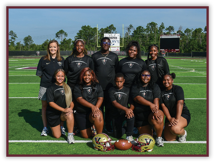 A group of adults and one child wearing matching black sports shirts pose on a football field with two helmets and a football placed in front of them. Description generated by AI