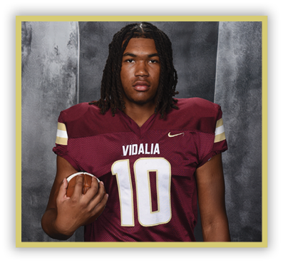 A person with shoulder-length dreadlocks wearing a maroon football jersey with the number 10 and the word Vidalia is holding a football. Description generated by AI