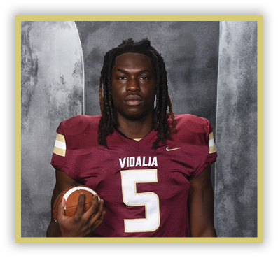A person with long dreadlocks wearing a maroon football jersey with the number five and the word Vidalia is holding a football against a gray textured background. Description generated by AI