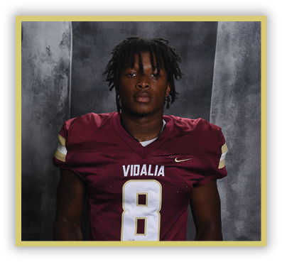 A person with short dreadlocks wearing a maroon football jersey with the word Vidalia and the number 8 stands against a dark gray backdrop. Description generated by AI