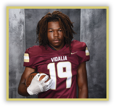A person with dreadlocks wearing a maroon football jersey with the number 19 and the word Vidalia is holding a football against a gray backdrop. Description generated by AI