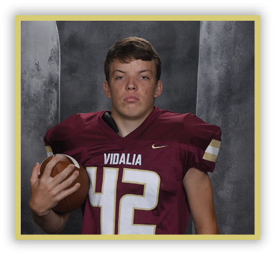 A person wearing a maroon football jersey with the number 42 and the word Vidalia is holding a football against a dark gray background. Description generated by AI