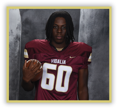 A person with shoulder-length dreadlocks wearing a maroon football jersey with the number 60 and the word Vidalia is holding a football against a gray backdrop. Description generated by AI