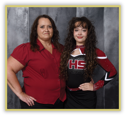 Two people stand side by side against a gray textured backdrop, one wearing a red button-up shirt and the other dressed in a black and red cheerleading uniform with long curly hair. Description generated by AI