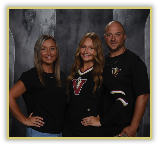 Three adults stand closely together smiling against a dark textured backdrop, with one wearing a black outfit featuring a large letter V and the others in casual black tops. Description generated by AI