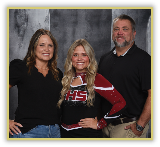 Three adults stand closely together smiling, with one wearing a cheerleading uniform and the other two dressed in casual clothing against a dark backdrop. Description generated by AI