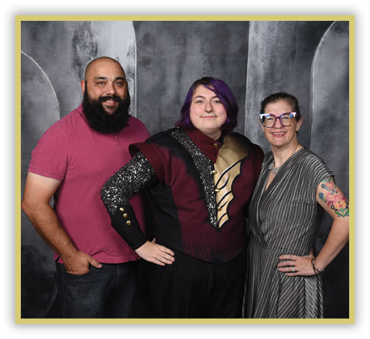 Three people stand closely together smiling, with one wearing a maroon and black costume with decorative details, another in a pink shirt, and the third in a striped dress with colorful tattoos on one arm and large glasses. Description generated by AI
