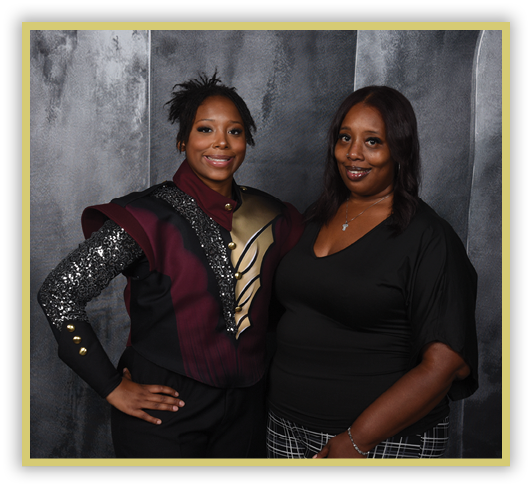 Two adults stand side by side against a dark textured background, one wearing a decorative costume with sequins and metallic accents and the other dressed in a black top and patterned pants. Description generated by AI