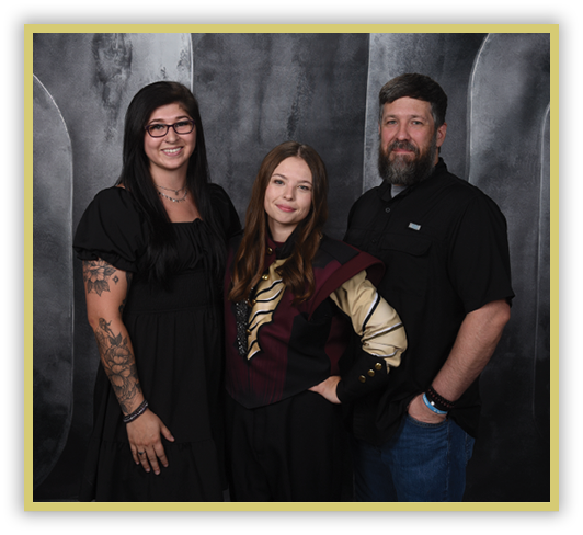 Three adults stand closely together against a dark textured backdrop, with one wearing glasses and a black dress showing tattoos, another in a maroon and beige patterned top, and the third in a black shirt and jeans smiling. Description generated by AI