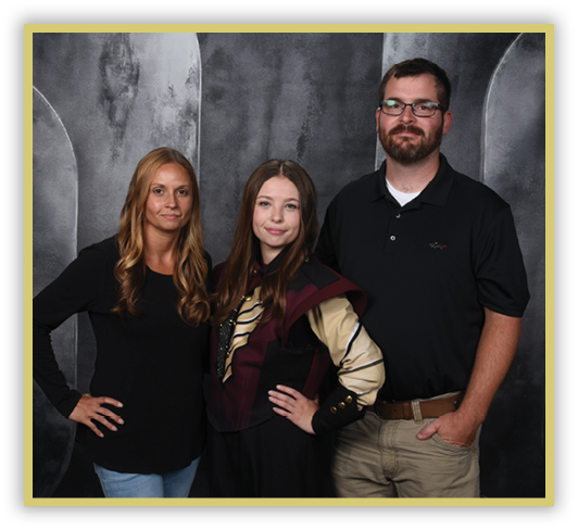 Three adults stand closely together against a dark textured backdrop, with one wearing a black shirt and glasses, another in a black long-sleeve top and light jeans, and the third dressed in a costume with a maroon and beige jacket. Description generated by AI