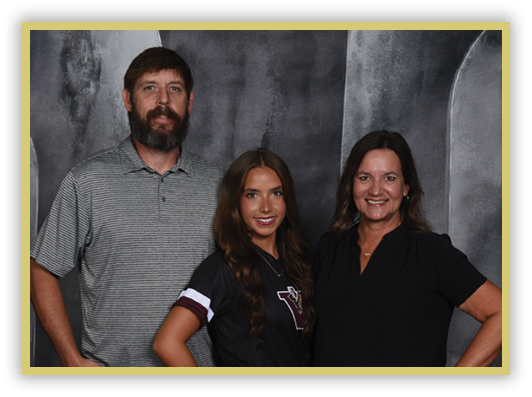 Three adults stand closely together smiling against a dark textured backdrop, with one wearing a striped polo shirt, another in a black sports jersey, and the third in a black blouse. Description generated by AI