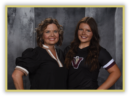 Two adults stand side by side smiling against a gray textured backdrop, one wearing a black blouse with puffed sleeves and the other wearing a black sports jersey with a “V" logo. Description generated by AI